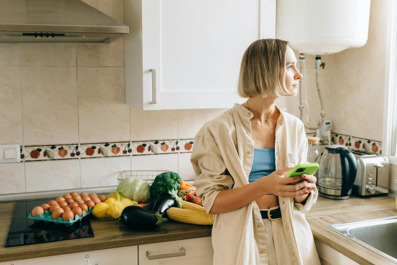Woman in a kitchen holding a green phone, looking out the window. On the counter are fresh vegetables and eggs. The mood is calm and contemplative.