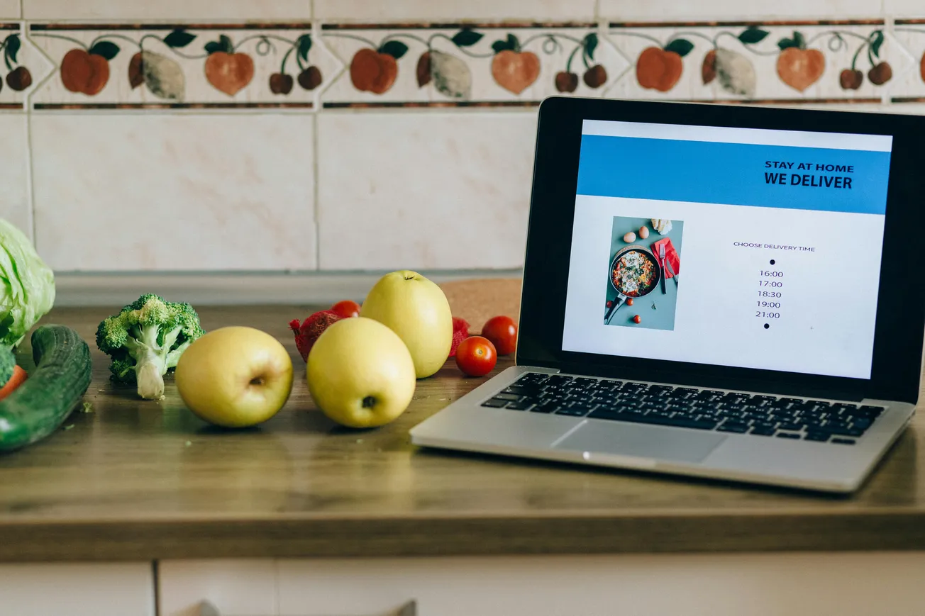 Laptop on a kitchen counter shows a food delivery ad. Nearby are apples, tomatoes, lettuce, broccoli, and a cucumber, suggesting freshness and health.