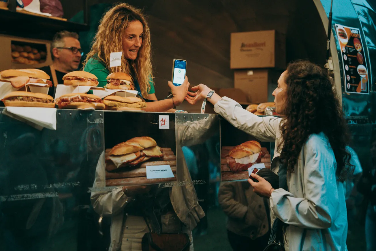 A woman at a food stall shows her phone to a vendor while smiling. Burgers line the counter, and the atmosphere is casual and vibrant.