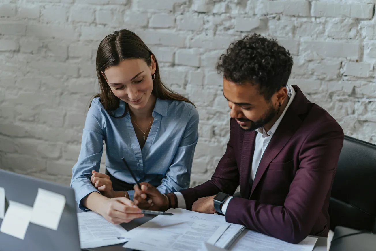 Two colleagues sit at a desk, smiling as they review documents. The man points at papers with a pen, while the woman attentively listens. They exude teamwork.