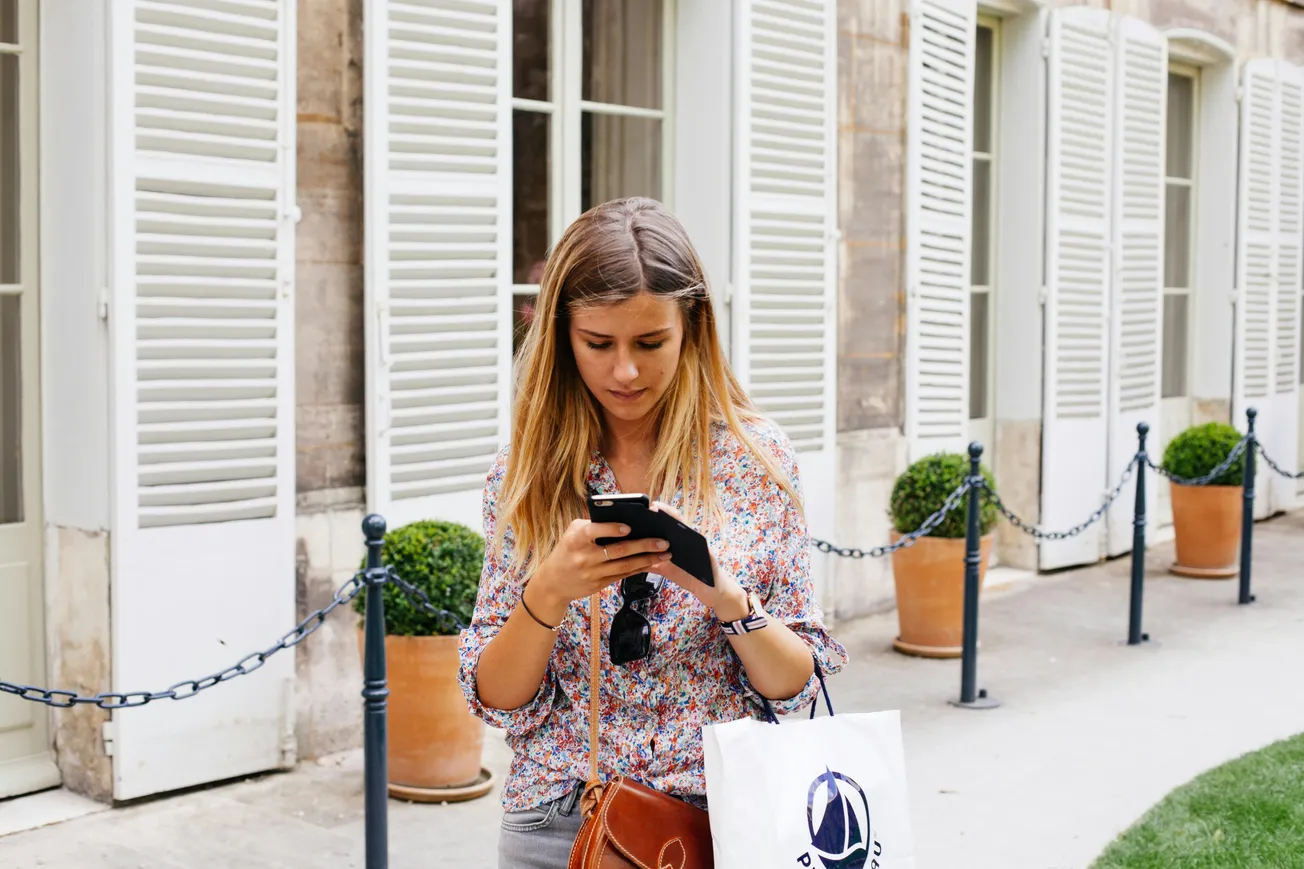 A woman with long hair, wearing a floral blouse, is focused on her smartphone while holding a shopping bag. Behind her are tall windows with shutters and potted plants.