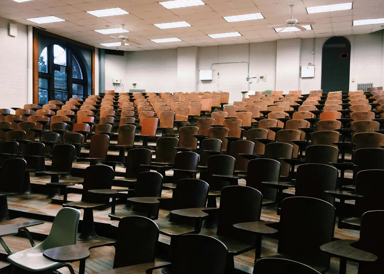 Empty lecture hall with rows of wooden desks and chairs, soft lighting overhead, and large arched windows on the side, conveying a sense of quiet and anticipation.