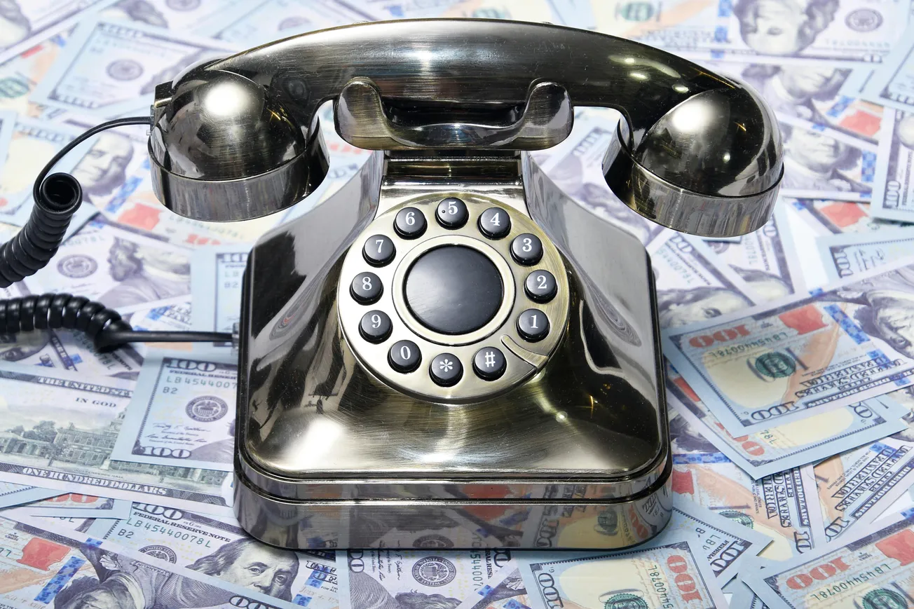 A shiny, vintage rotary phone is placed on a spread of one hundred dollar bills. The image conveys a sense of wealth and retro communication.