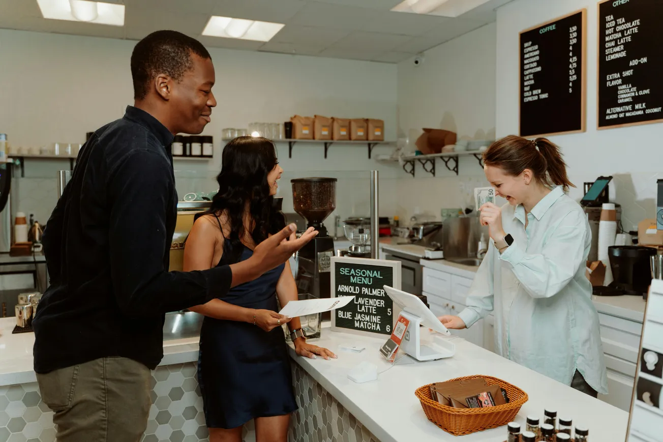 Two customers, a man and a woman, happily order at a bright café counter from a smiling barista. The menu features seasonal drinks. Cozy atmosphere.