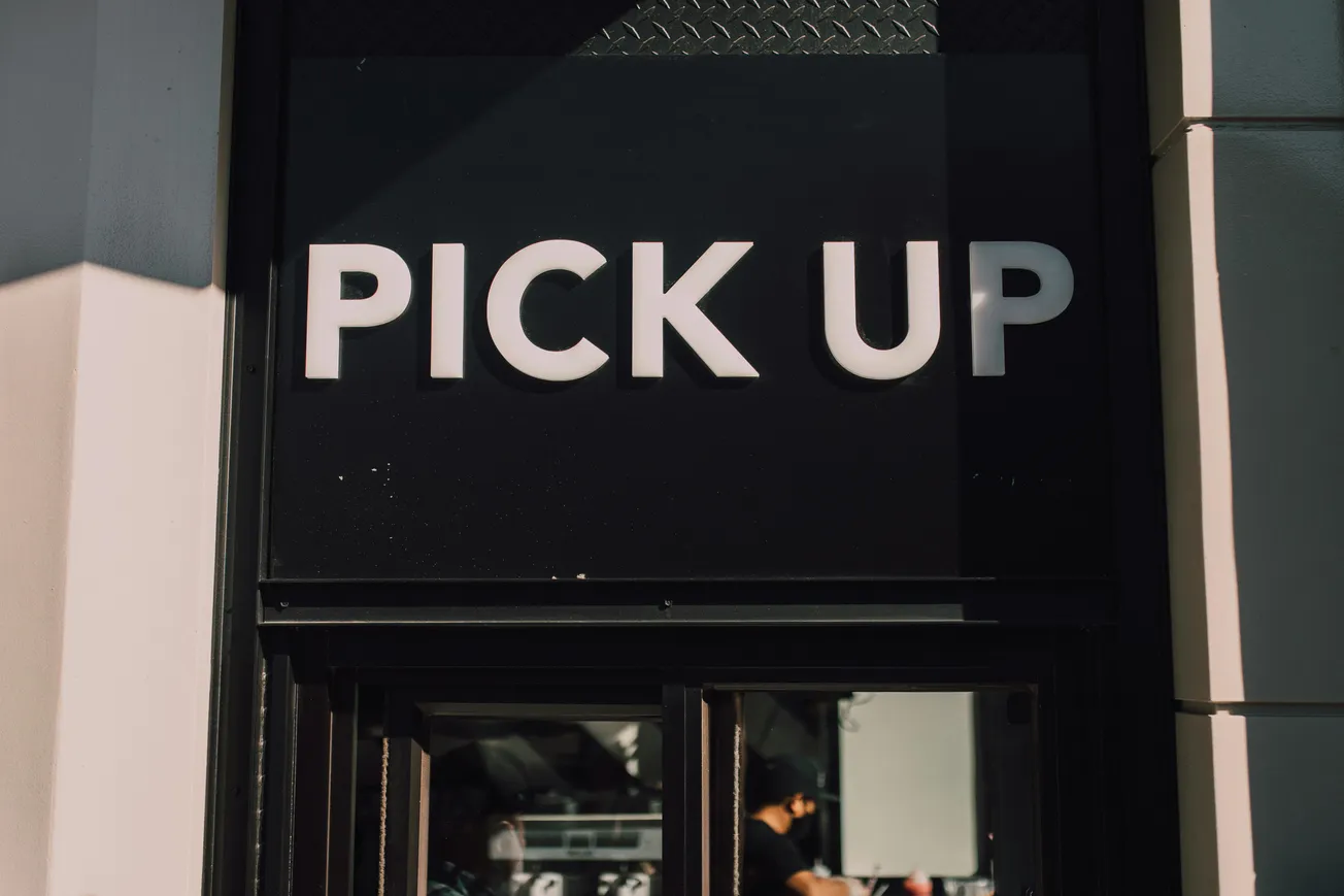 Sign reading "PICK UP" in bold white letters against a black background, with a small window below. Bright sunlight creates strong shadows.