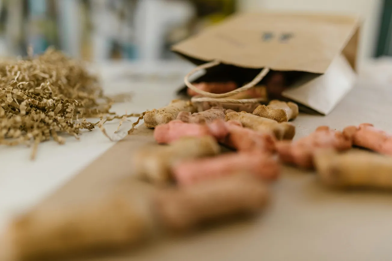 A paper bag spills colorful dog treats onto a surface, surrounded by shredded packing material. The scene feels playful and casual.