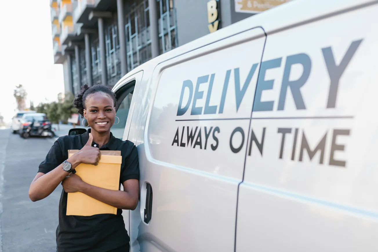 Smiling delivery worker holding an envelope stands beside a van labeled "DELIVERY ALWAYS ON TIME" on an urban street, conveying reliability.