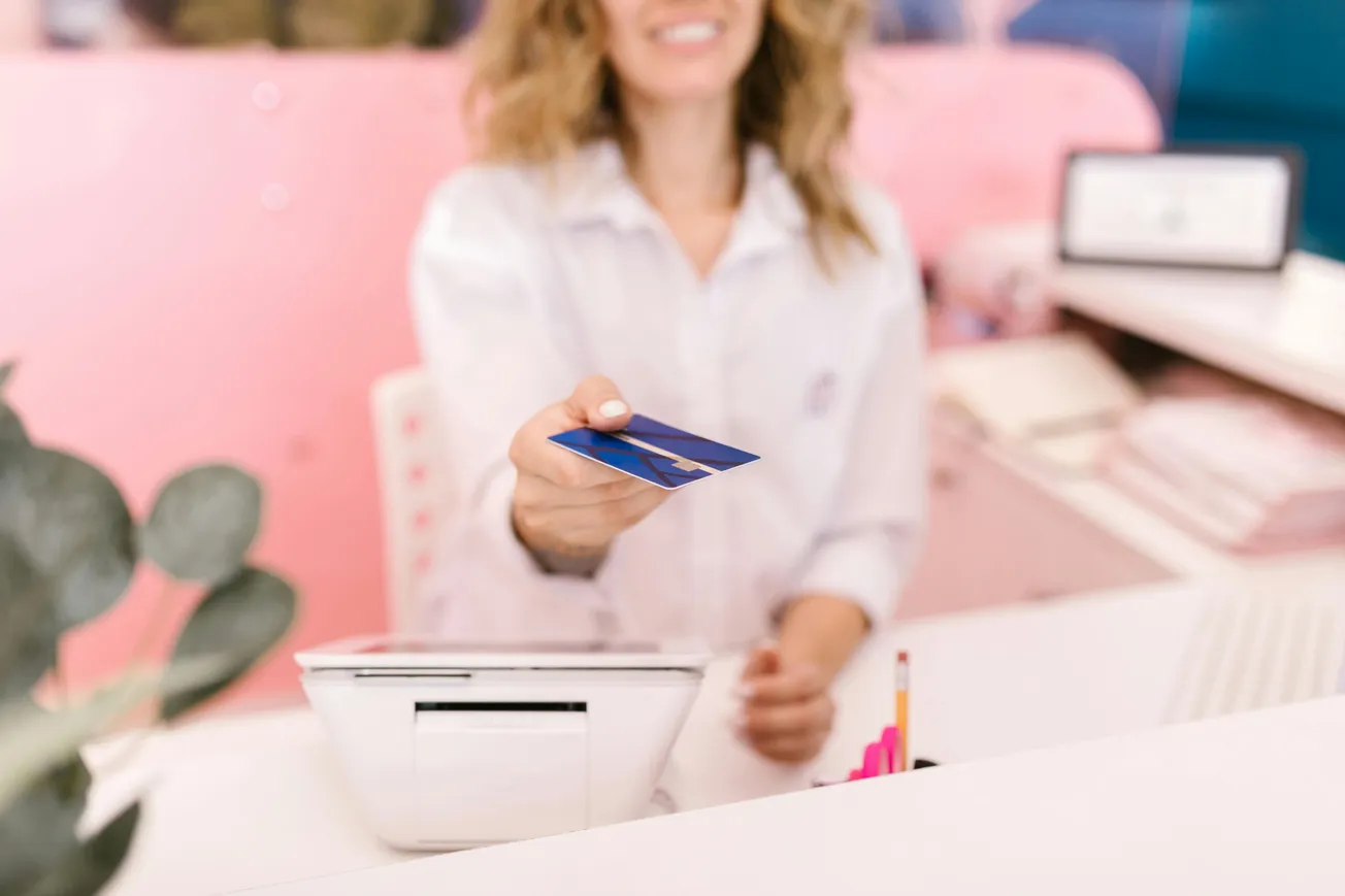 Woman smiling, handing a blue credit card across a white counter, with a payment terminal in the foreground. The background is a soft pink.