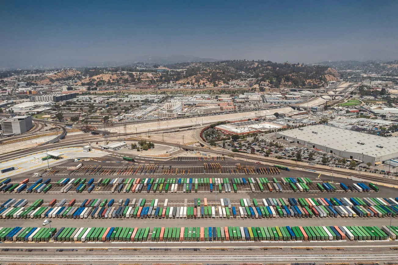 Aerial view of a busy freight yard with colorful cargo containers lined up in rows. In the background, urban infrastructure and hilly terrain are visible.