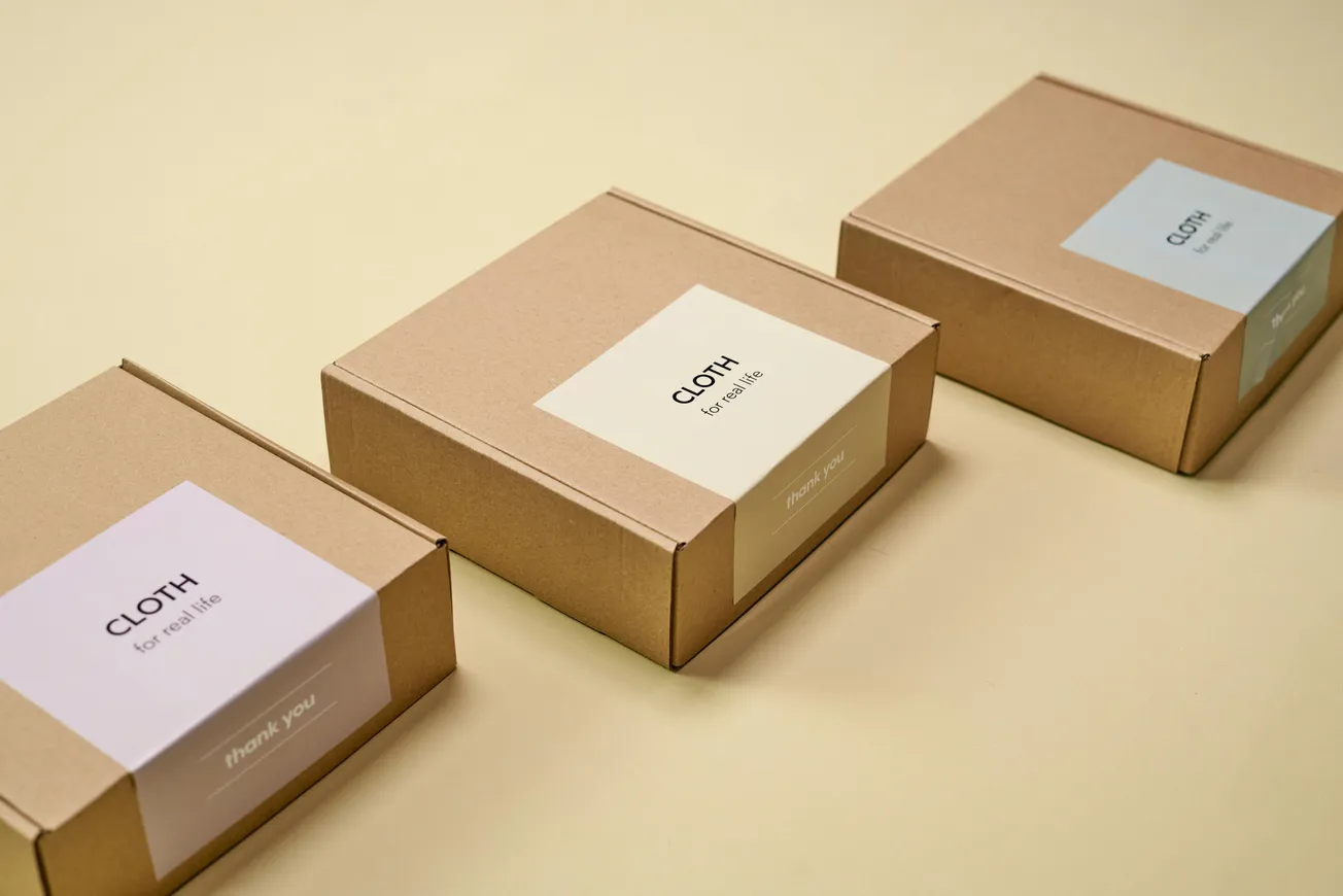 Three brown cardboard boxes are displayed on a beige surface, each labeled "CLOTH." The mood is minimalistic and organized.
