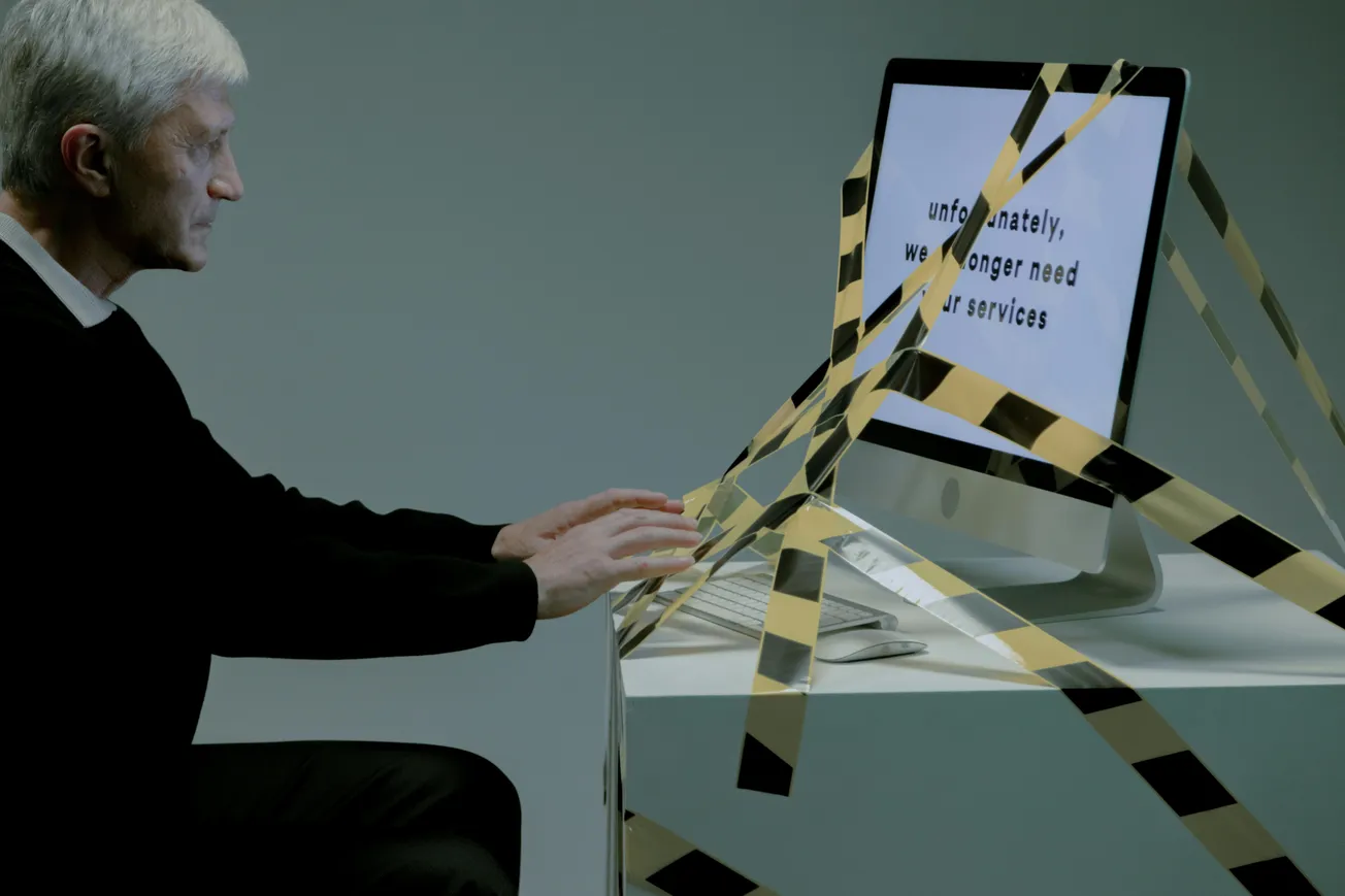 An older man sitting at a desk with his hands near a computer wrapped in caution tape. The screen displays a message about no longer needing services, conveying job loss.