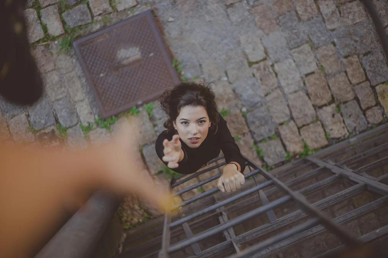 Woman climbing a metal ladder, viewed from above, reaching towards an outstretched hand. Background shows cobblestone pavement, suggesting urgency.