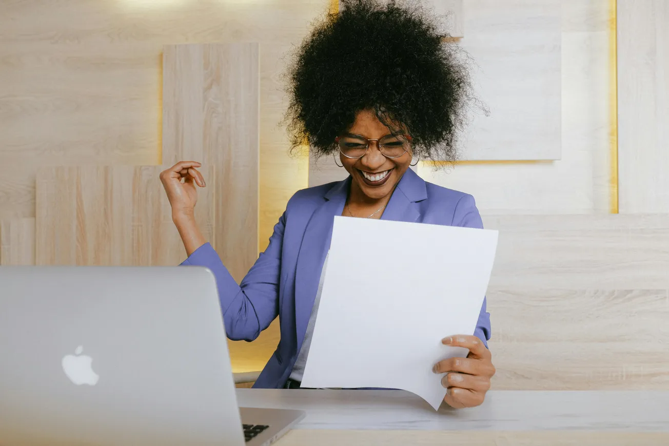 A woman with curly hair, wearing glasses and a blue blazer, joyfully reads a paper while sitting at a desk with a laptop, expressing excitement.