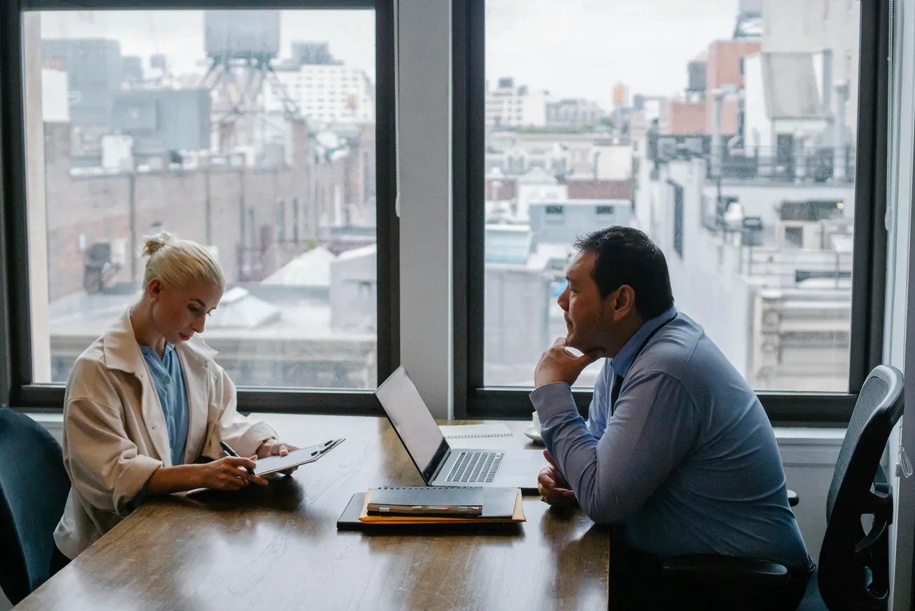 Two professionals are in a meeting room with large windows overlooking a cityscape. One is writing on a clipboard, and the other is focused on a laptop.