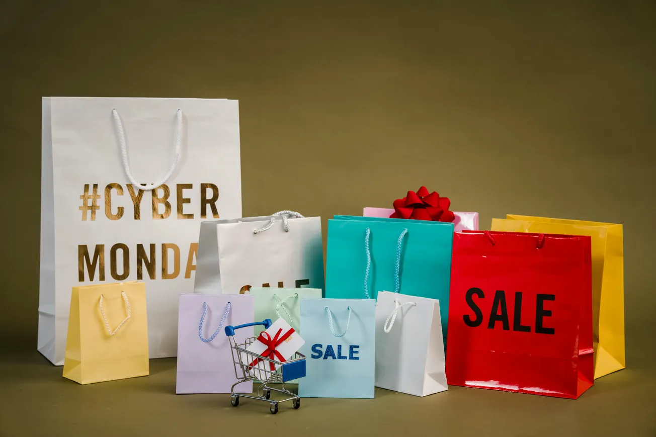 Shopping bags labeled “Cyber Monday” and “Sale” in various colors with a miniature cart and red bow, suggesting festive holiday shopping deals.