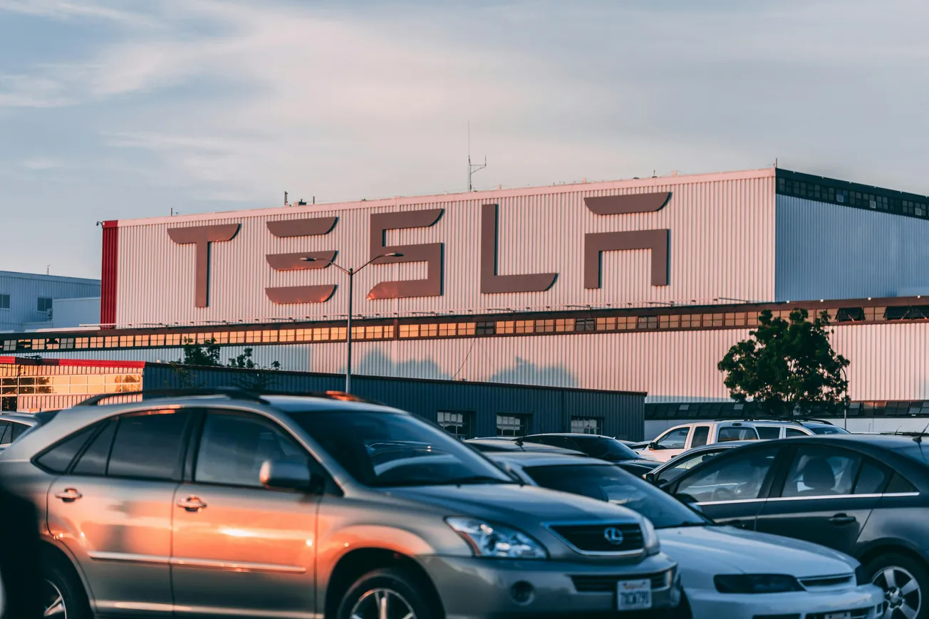 Parking lot at sunset with various cars, in front of a large building labeled "TESLA" in bold letters. A tree and light pole are visible.