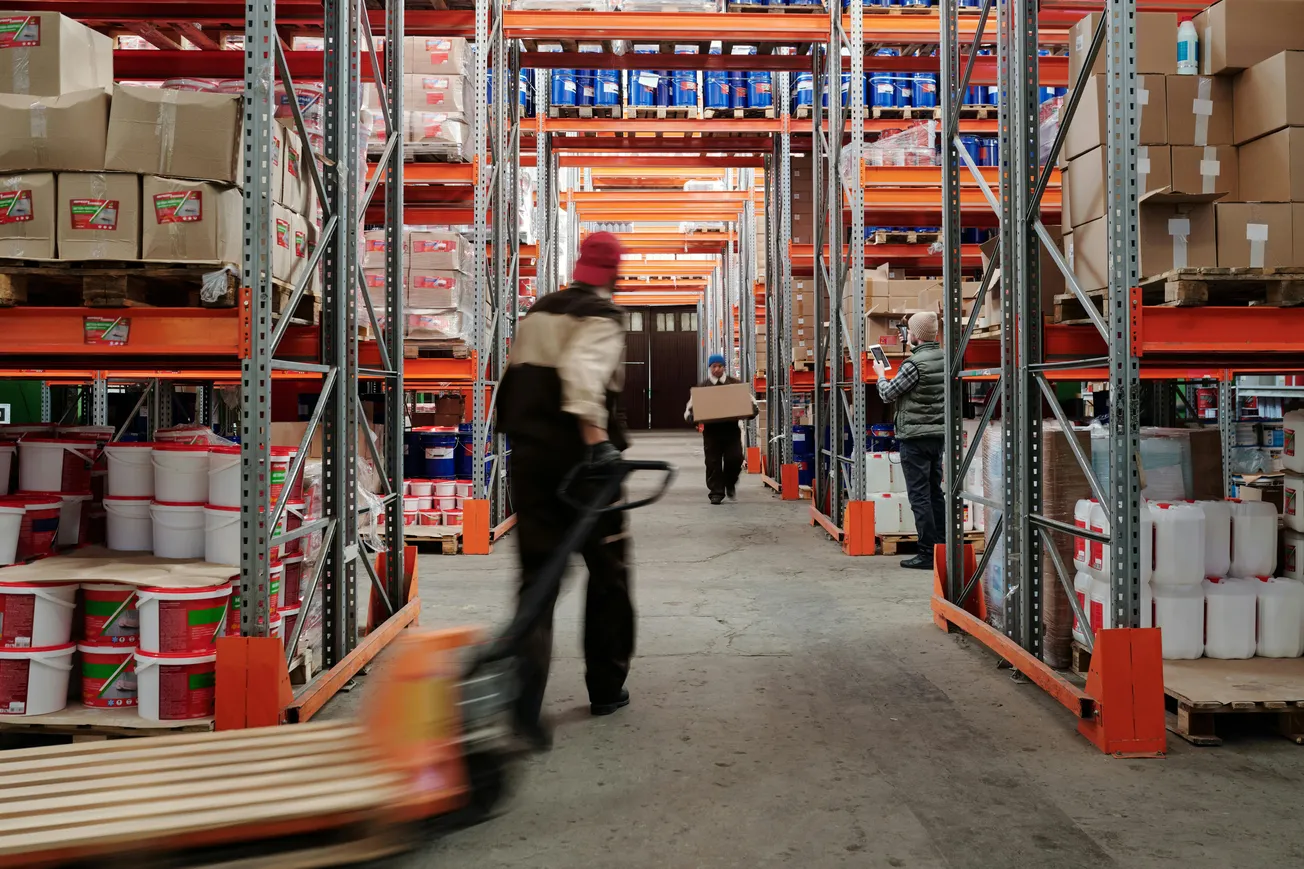 Warehouse scene with three workers among shelves stacked with boxes and containers. One pushes a pallet jack, another carries a box. Busy, organized atmosphere.