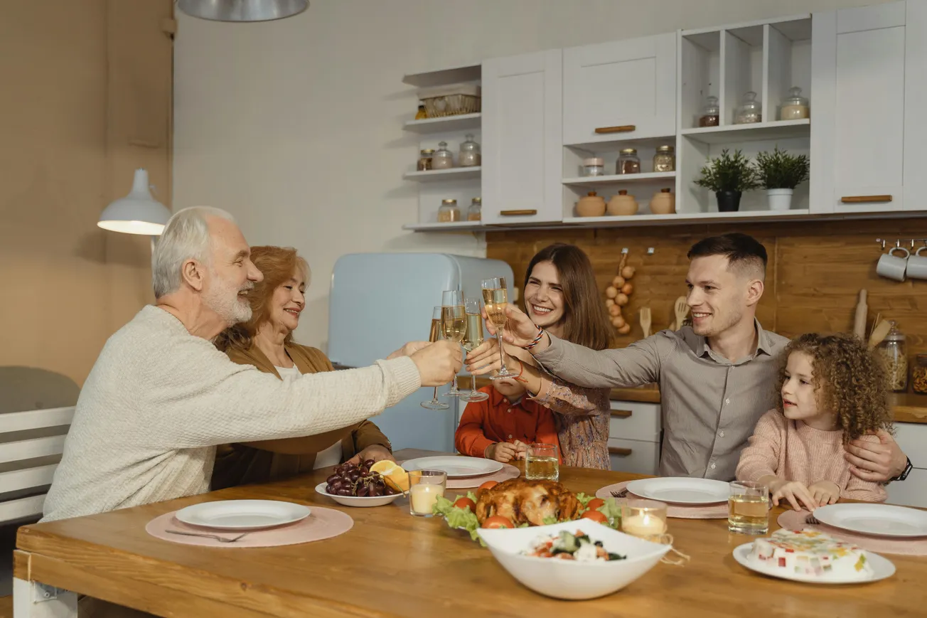 Family gathering in a cozy kitchen, raising glasses in a toast. A table is set with food, evoking warmth and celebration. Smiling faces convey joy.