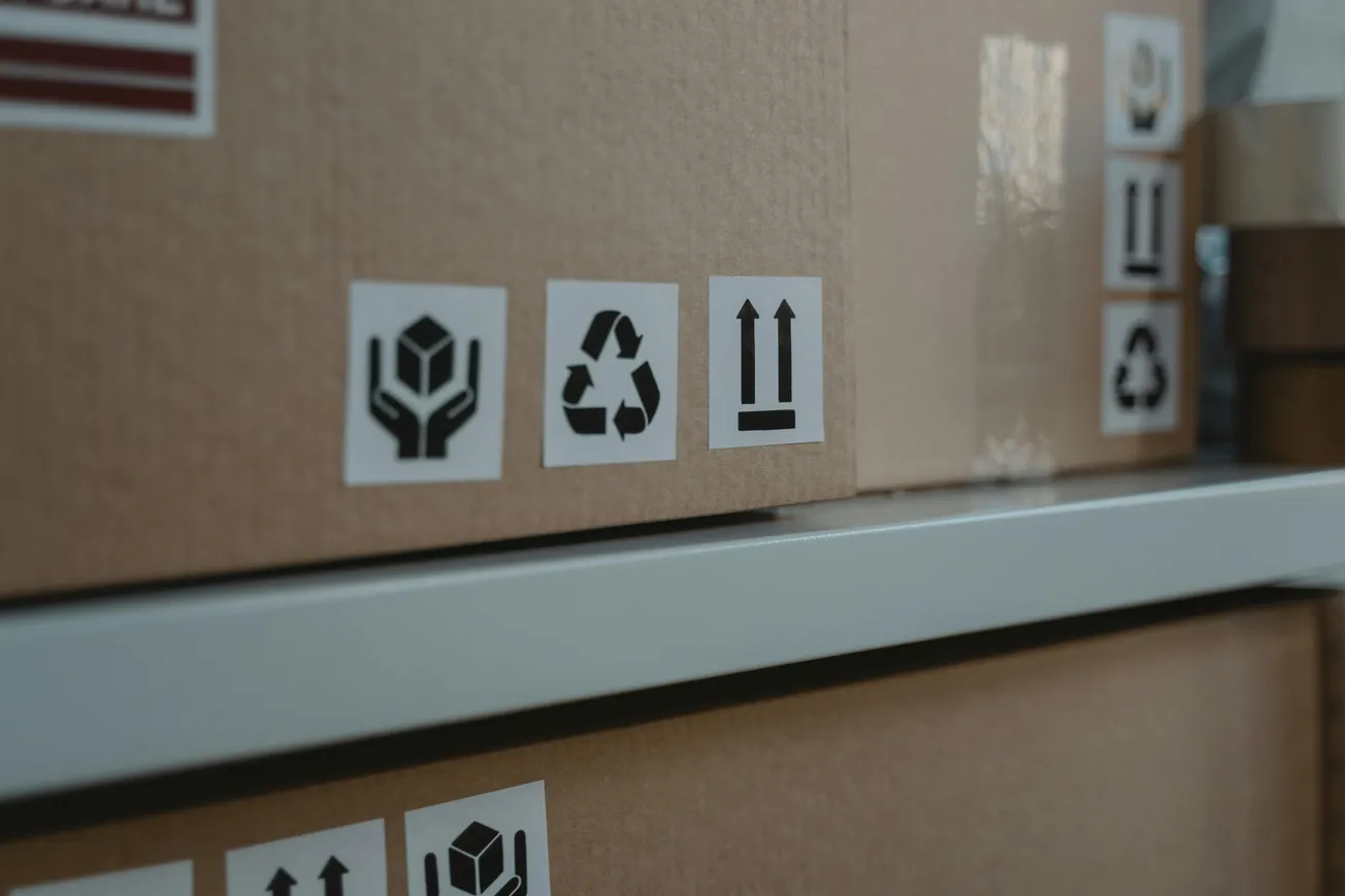 Brown cardboard boxes on a shelf, featuring handling symbols: fragile, recycling, and arrows indicating correct upright position. Represents packaging and shipping.