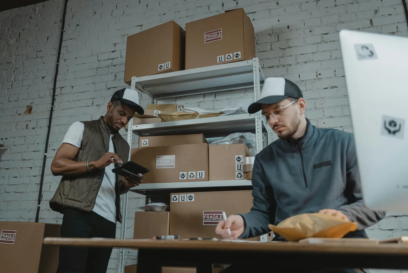 Two workers in a warehouse are organizing packages. One inspects a tablet, the other writes notes. Cardboard boxes labeled "Fragile" are stacked behind.