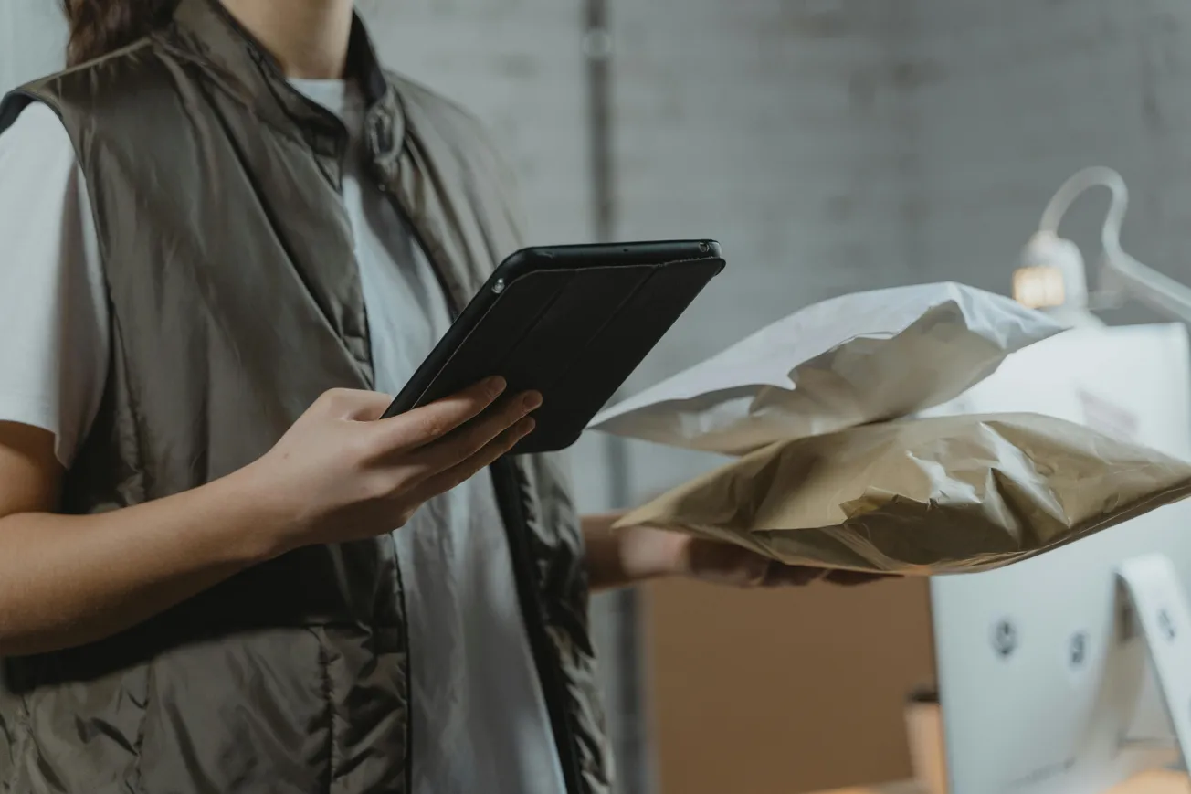 A person in a brown vest holds packages and a tablet in a dimly lit office. The scene suggests a busy, focused atmosphere with an emphasis on logistics.