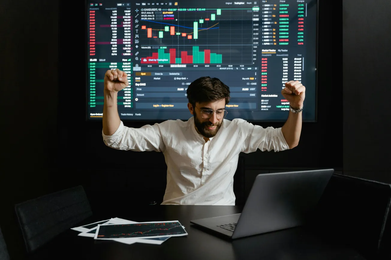 A man in a white shirt celebrates at a desk with a laptop, arms raised in triumph. A large screen behind shows colorful stock market charts and data.