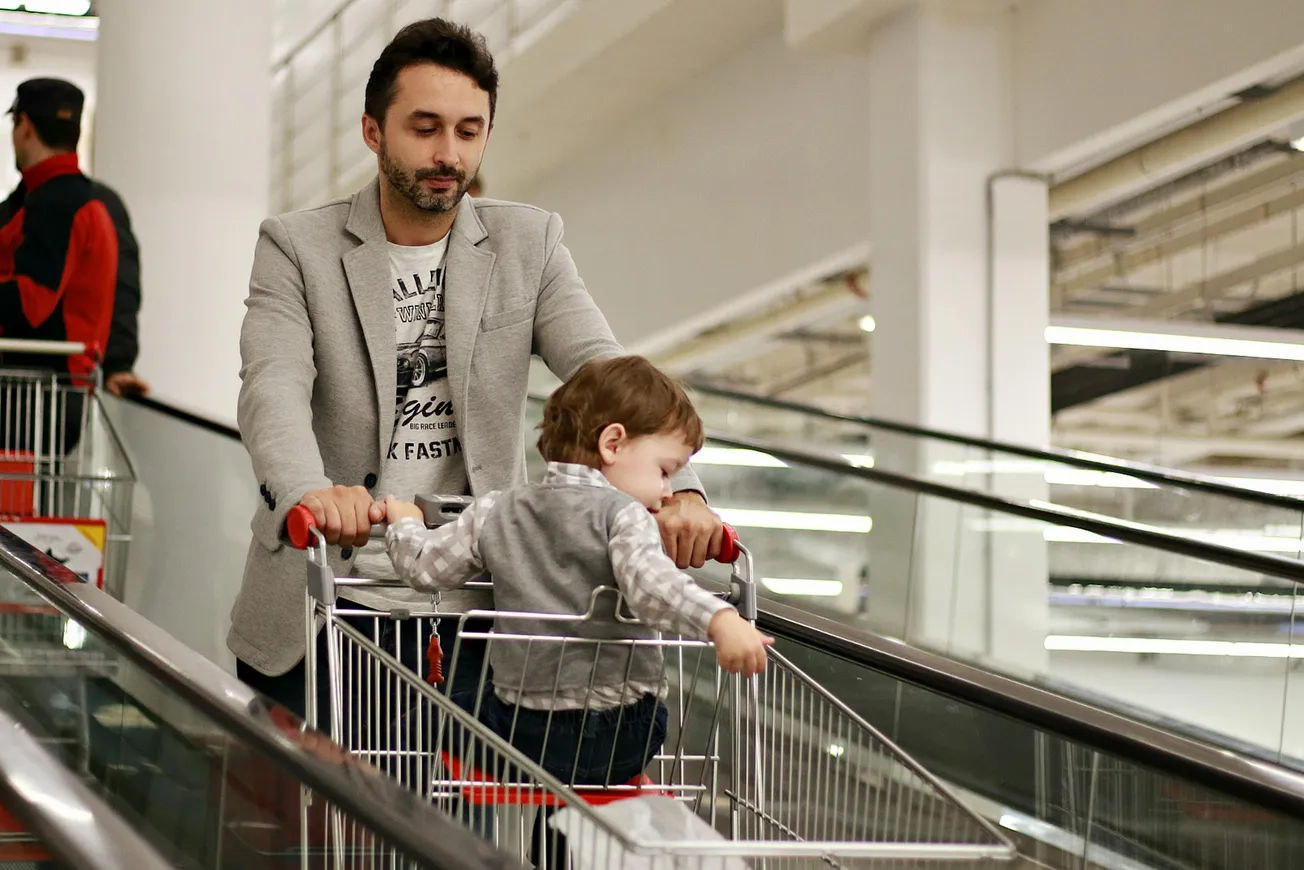 Man in a gray blazer pushes a young child in a shopping cart on a store's moving walkway. The setting is bright and modern, conveying everyday life.