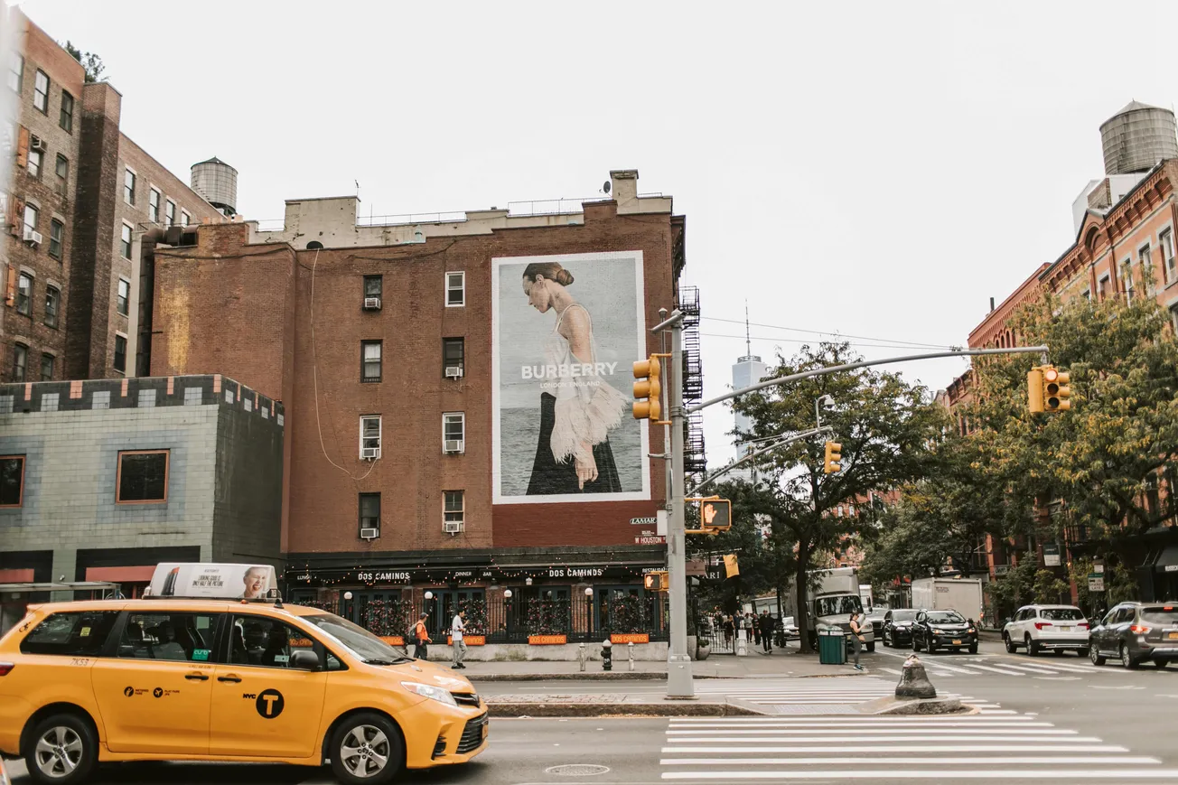A large Burberry ad featuring a woman in profile is displayed on a red brick building. A yellow taxi and pedestrians are nearby. Urban, bustling vibe.