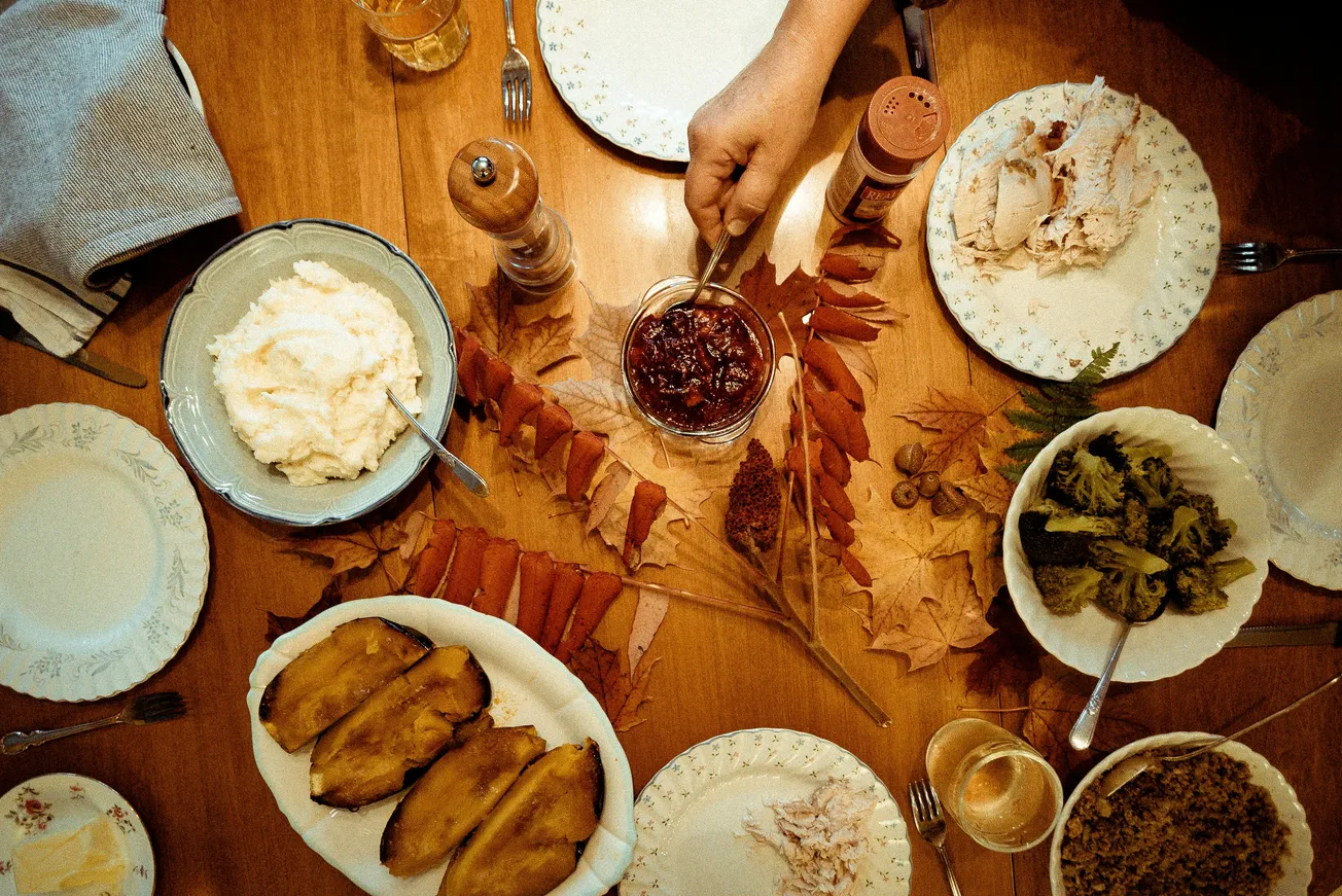 A Thanksgiving table filled with mashed potatoes, sliced turkey, cranberry sauce, roasted squash, and greens, adorned with autumn leaves.