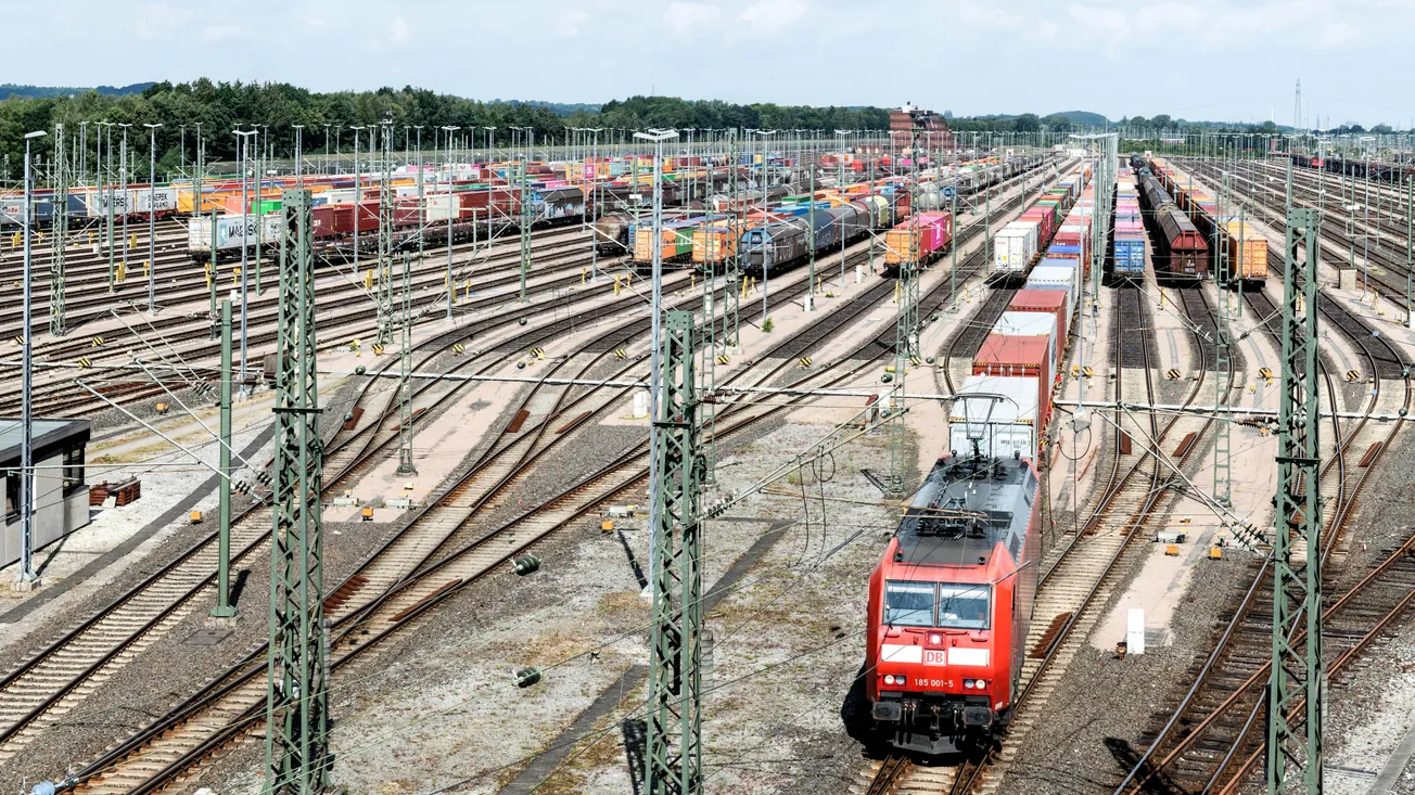 A vast railway freight yard with numerous tracks filled with colorful shipping containers. A red locomotive moves forward, conveying busy activity and industrial efficiency.