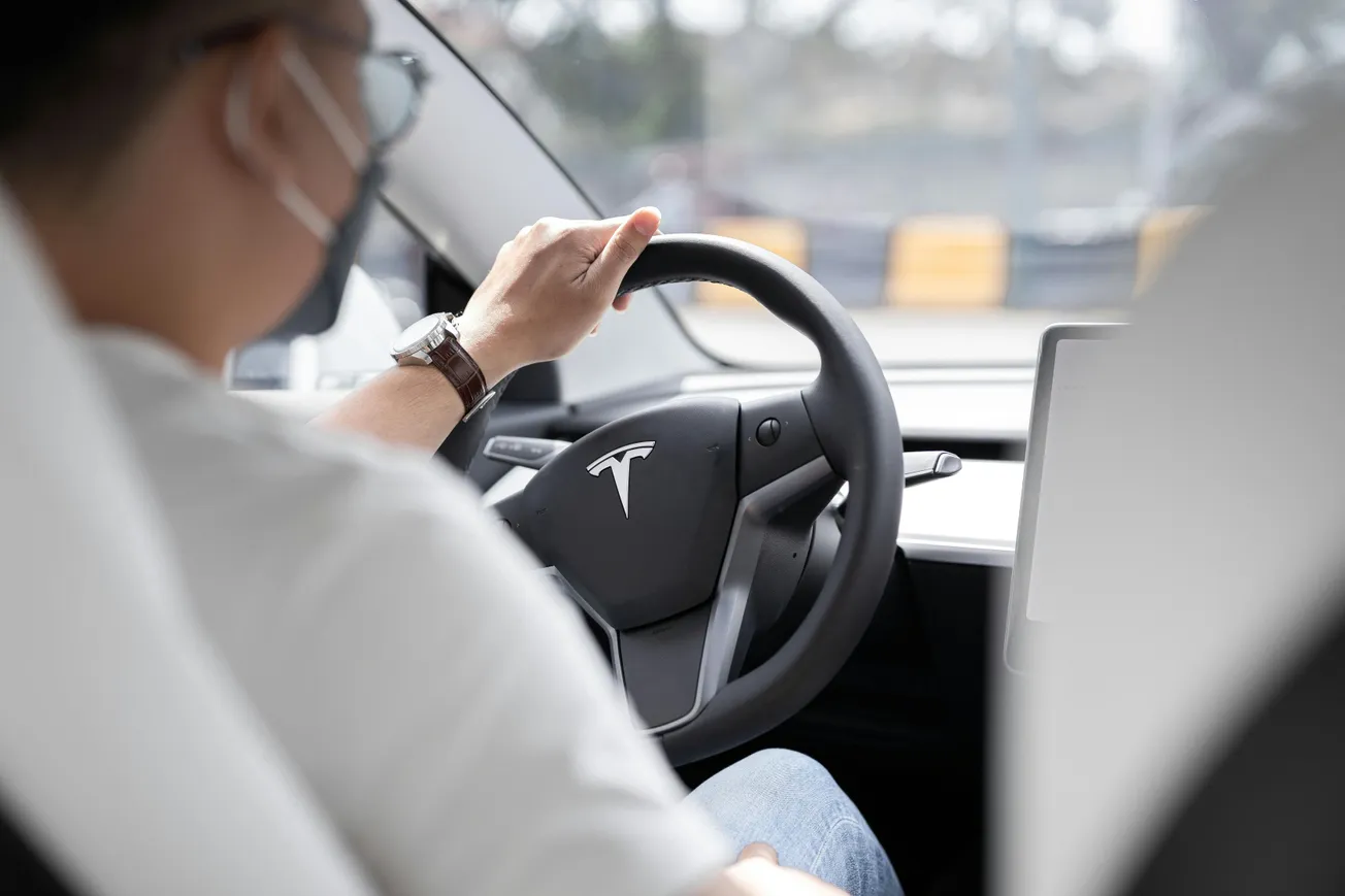 A person wearing a mask is driving a Tesla, focusing on the road. The Tesla logo is visible on the steering wheel, conveying a modern and safe atmosphere.