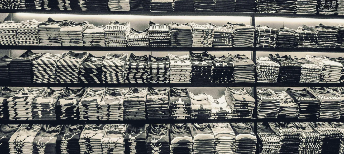 Stacks of folded, patterned shirts in various geometric designs fill several shelves in a store. The monochrome tones suggest an orderly, calm atmosphere.