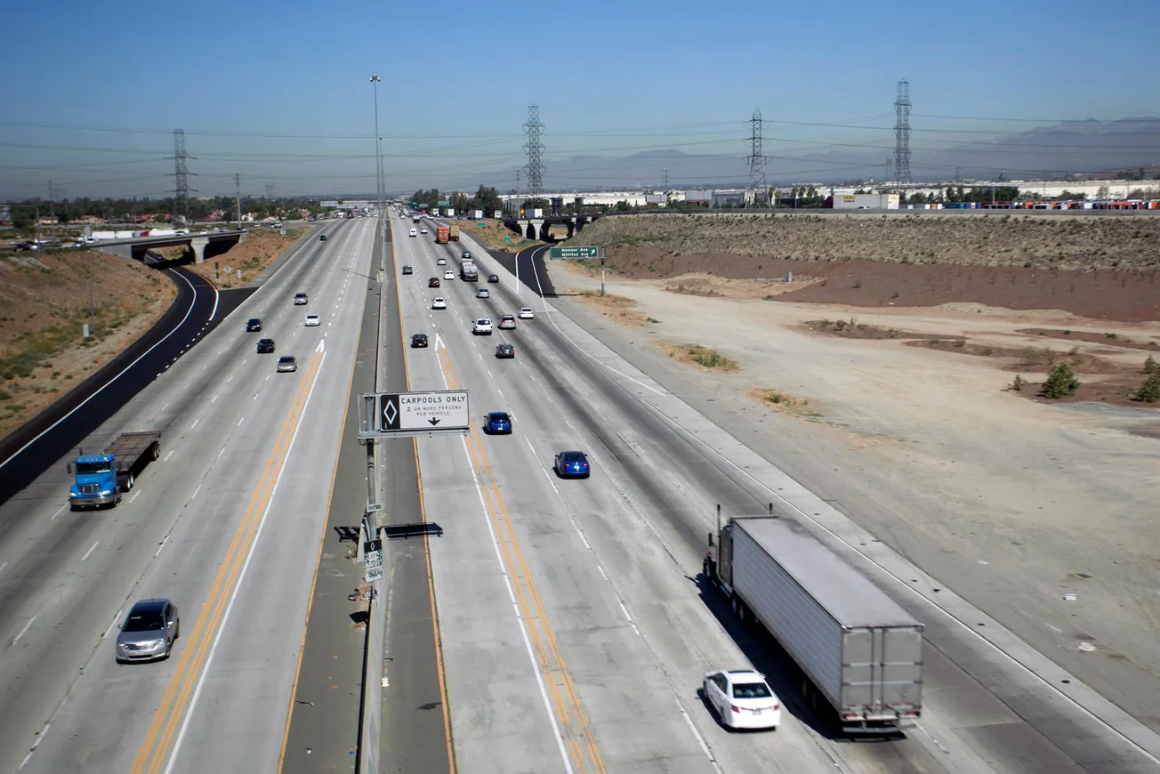Wide highway in daylight, sparse traffic with trucks and cars, bordered by desert landscape. Clear sky, industrial buildings in the distance. Calm atmosphere.