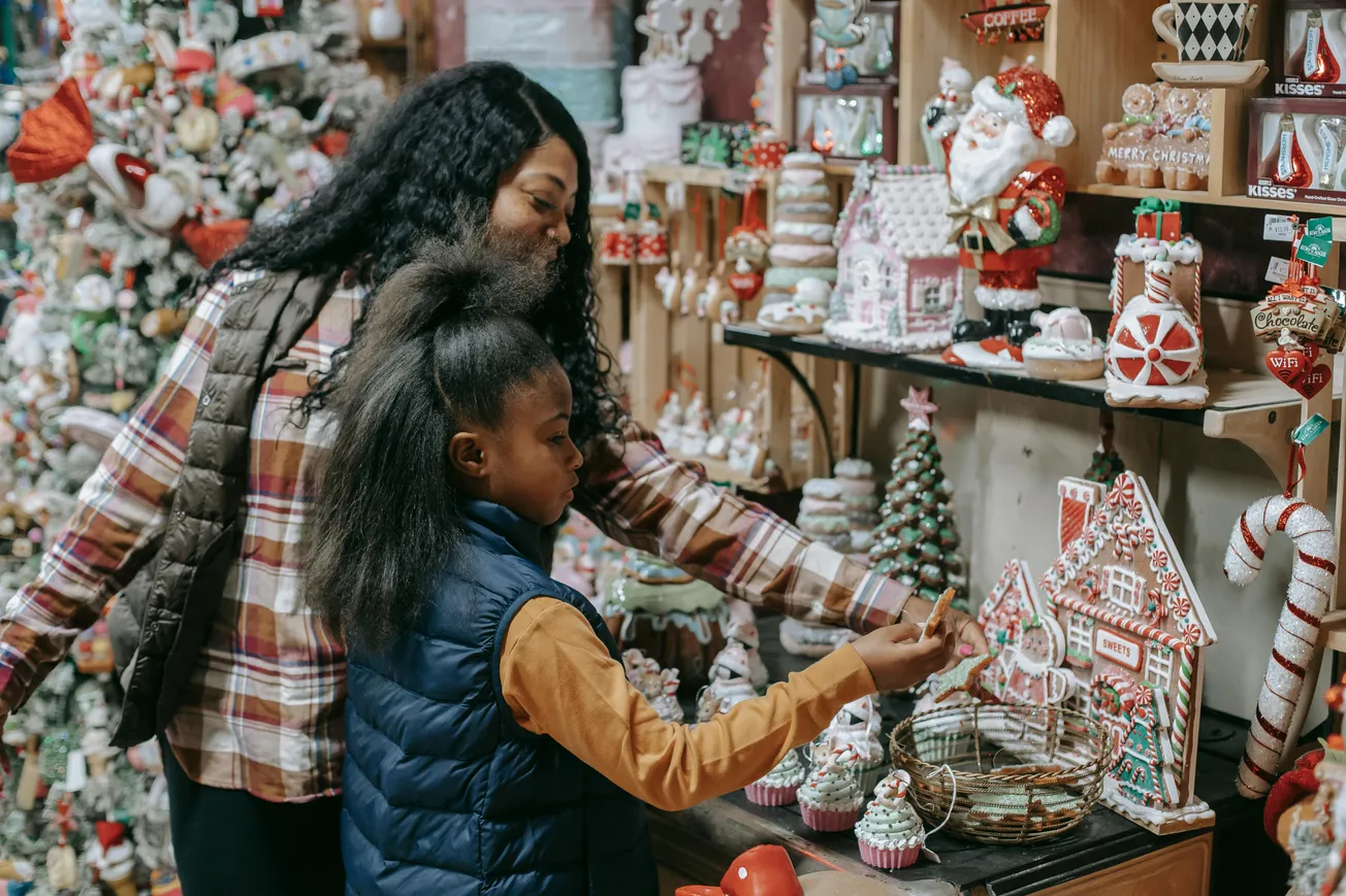 A mother and daughter admire holiday decorations in a store. The shelves are filled with festive items like gingerbread houses and Santa figurines, evoking a joyful atmosphere.