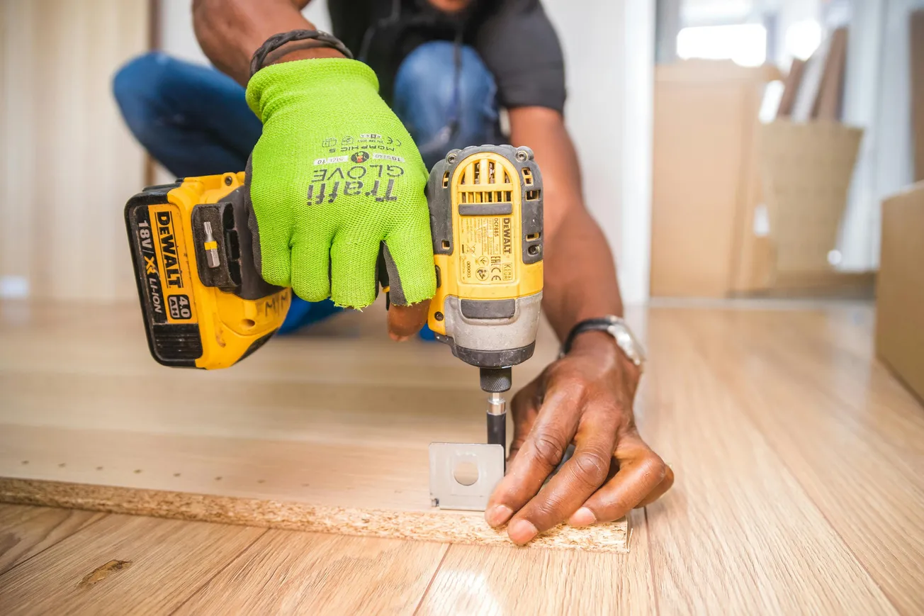 Person assembling furniture using a yellow drill and holding a bracket. Bright green glove on one hand, with a focused, industrious atmosphere.