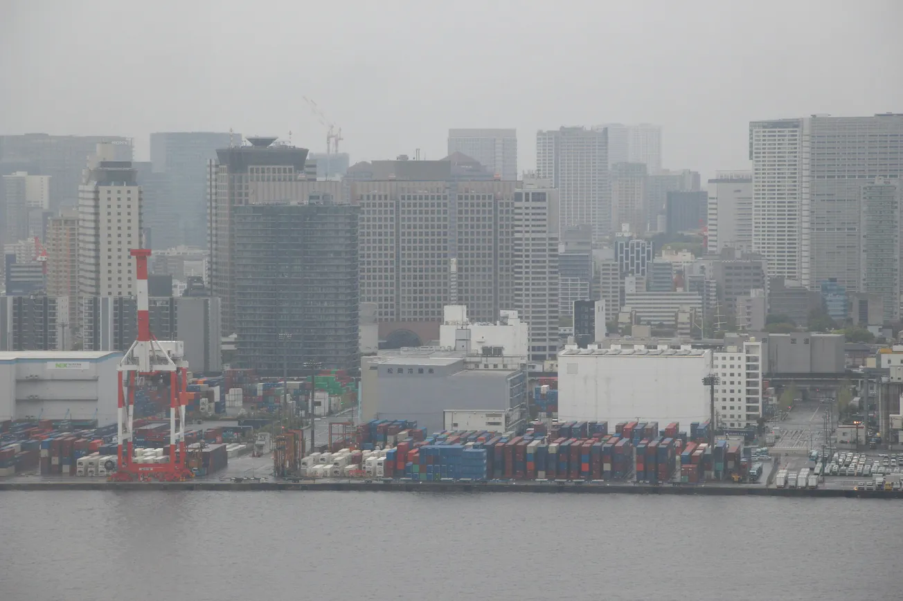 A cloudy city skyline with numerous high-rise buildings in the distance. In the foreground, a bustling port features stacked shipping containers and a crane.