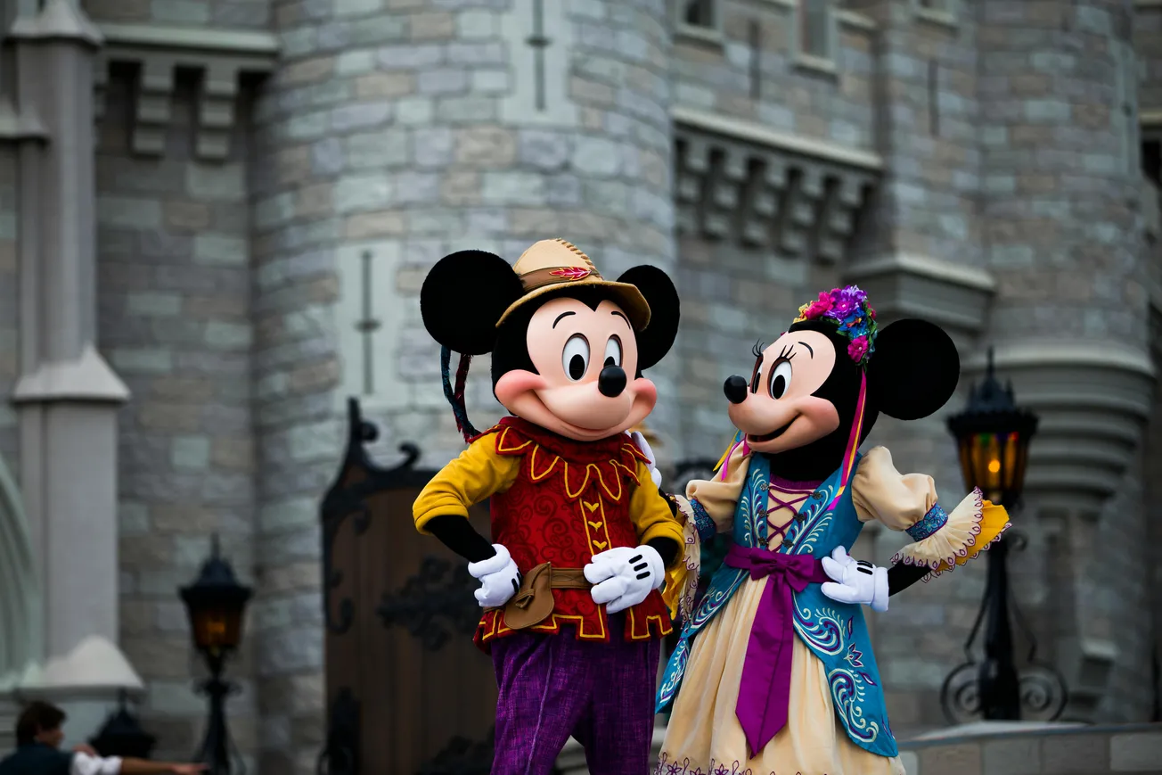 Mickey and Minnie Mouse in colorful, festive outfits, stand smiling outside a stone castle. The scene is lively, celebrating and joyful.