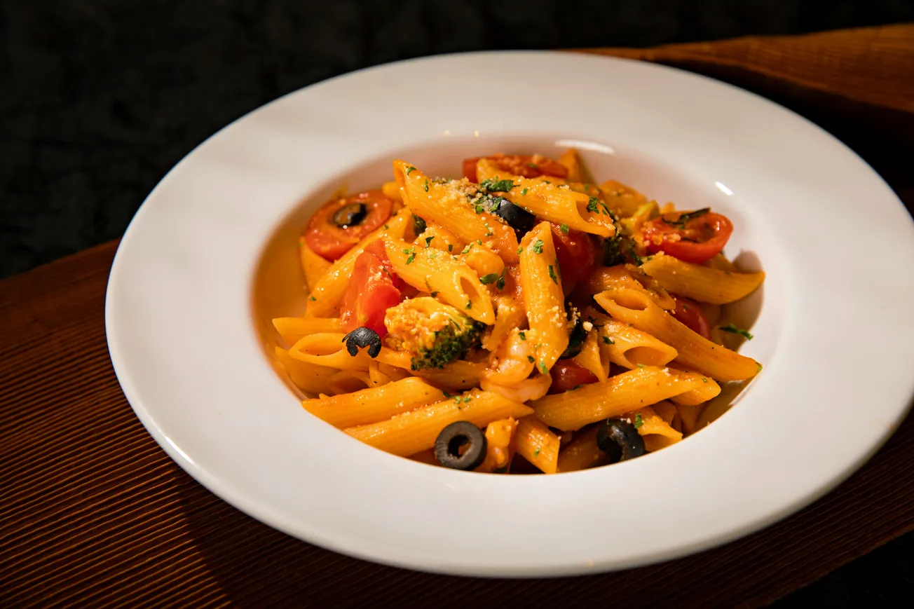 A close-up of a vibrant penne pasta dish garnished with cherry tomatoes, olives, and herbs on a white plate. The setting is warm and inviting.