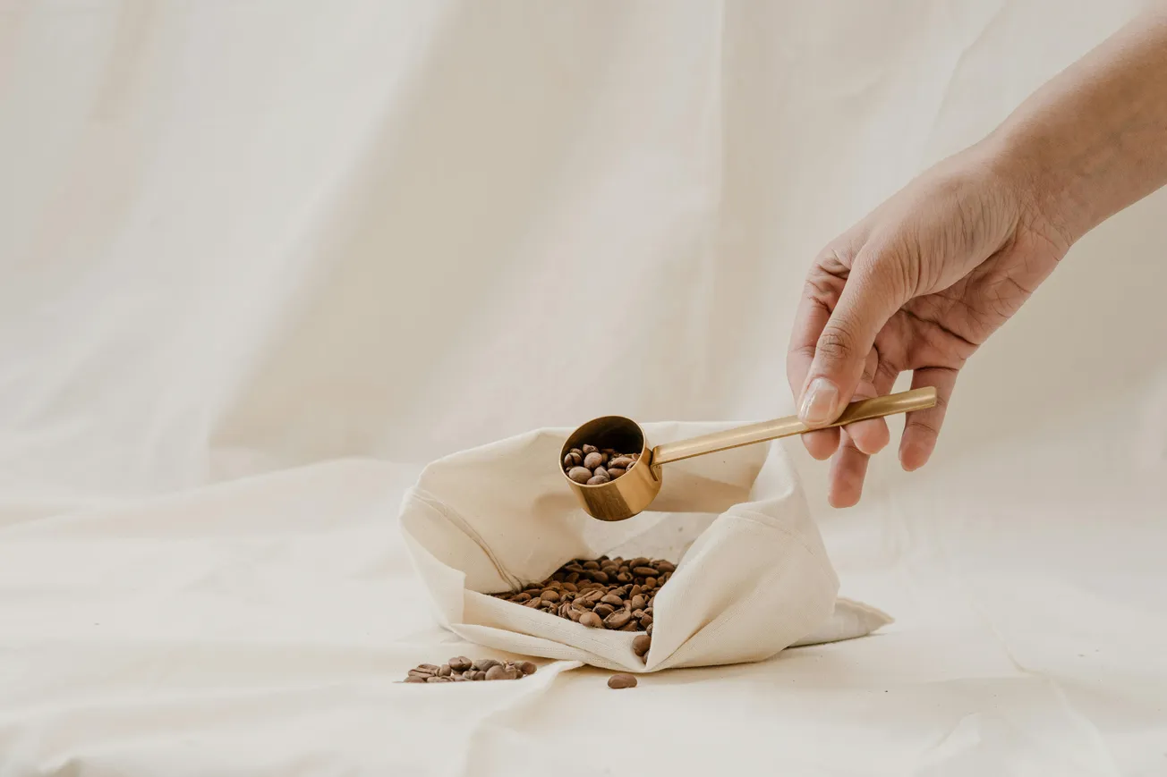 A hand holds a brass scoop above a cloth bag filled with coffee beans on a soft, cream-colored background, conveying a calm and serene tone.