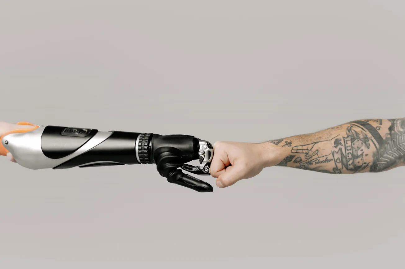 A tattooed human arm and a robotic prosthetic arm fist bump against a neutral background, symbolizing unity and technological integration.