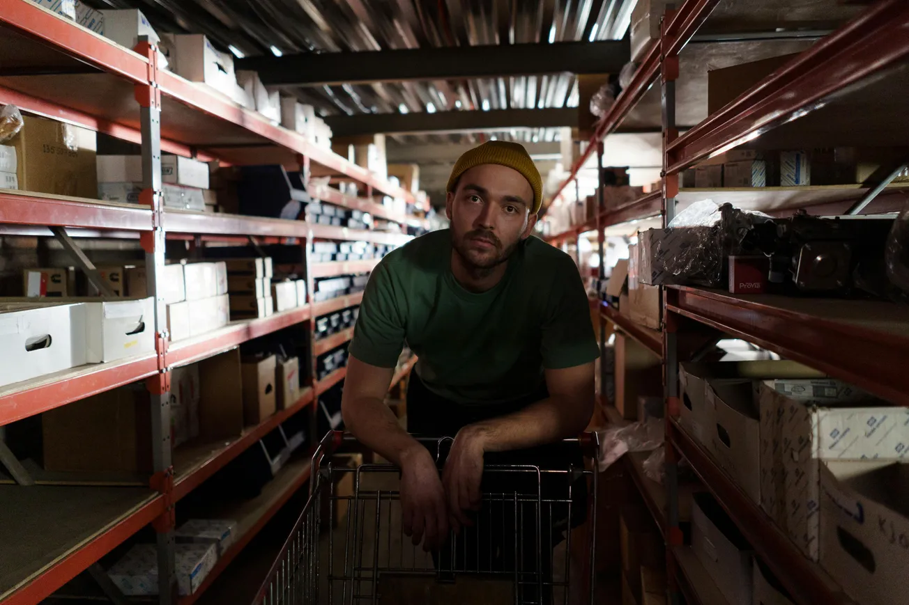 Man in green shirt and yellow beanie leans on shopping cart in dimly lit warehouse aisle. Shelves with boxes line both sides, creating a focused, contemplative mood.