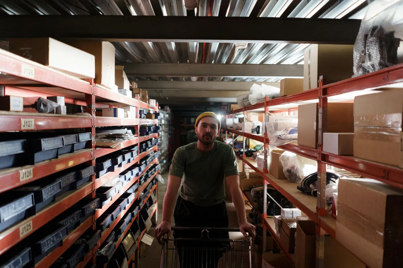 A person pushes a shopping cart down a dimly lit warehouse aisle, flanked by shelves filled with boxes and items. The atmosphere feels focused and industrious.
