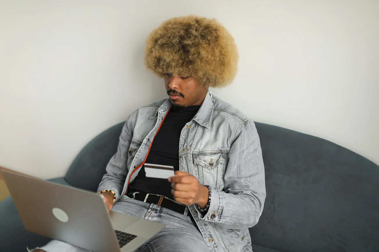 A person with curly hair sits on a couch, wearing a denim jacket, and focused on a laptop while holding a credit card.