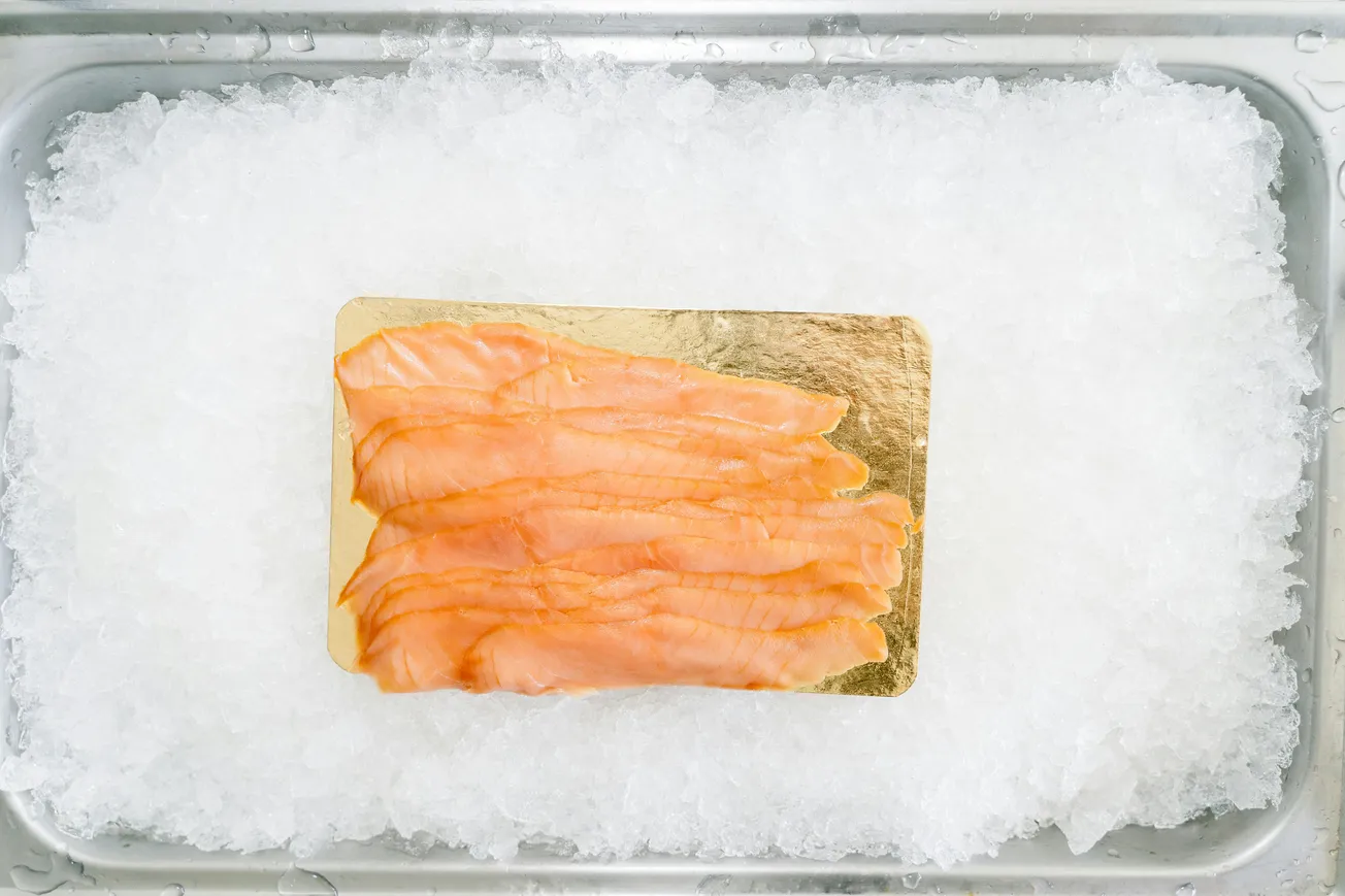 Slices of smoked salmon arranged on a gold-colored board resting on a bed of crushed ice in a metal tray, creating a fresh, chilled presentation.