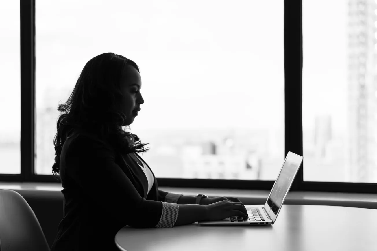 A woman sits at a table near a large window, typing on a laptop. The black-and-white image conveys a focused and professional atmosphere.