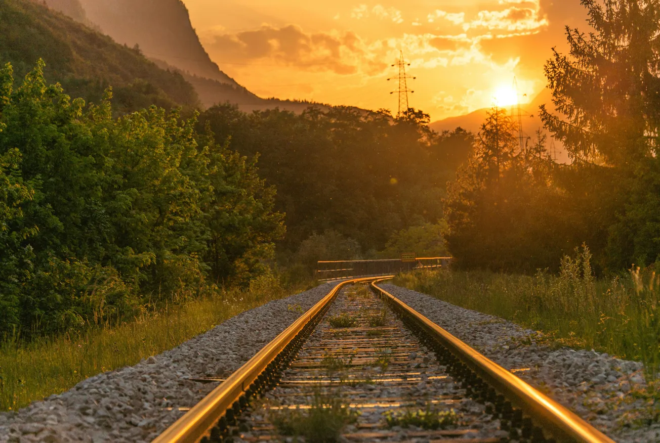 Sunset scene with train tracks leading into the distance, surrounded by lush trees and hills. The warm glow of the sun casts a peaceful, golden light.