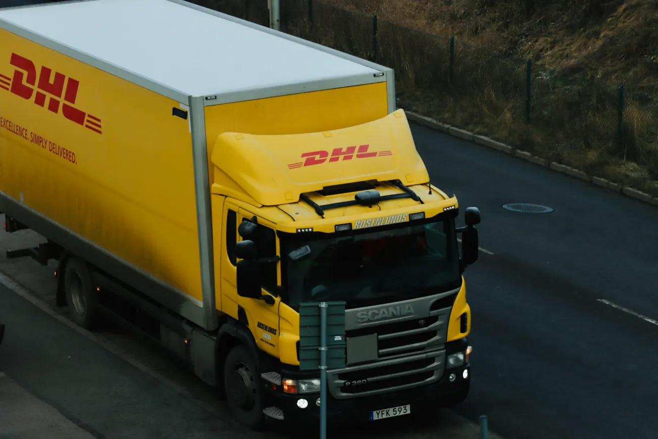 A yellow DHL delivery truck travels on a wet, empty road. Its logo is prominently displayed on a clean, boxy cargo section. Scene appears overcast.