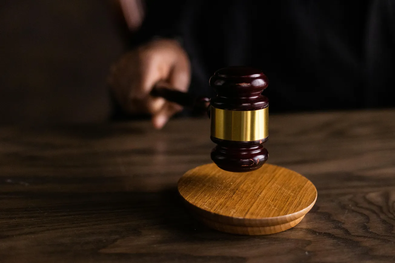 A judge's hand grips a wooden gavel poised above a round sound block on a wooden table, symbolizing law, justice, and authority in a courtroom setting.