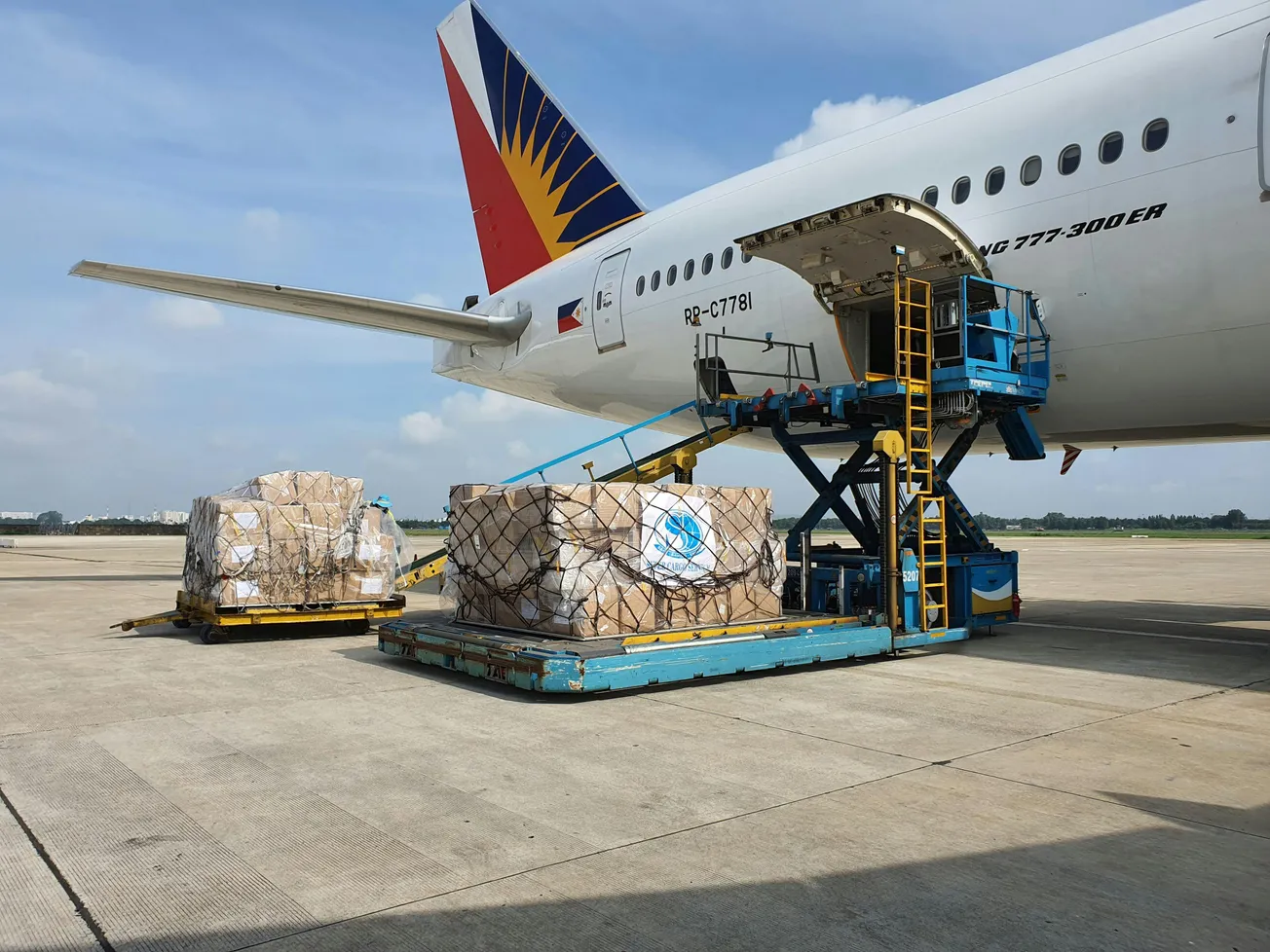 Cargo being loaded onto a large airliner via a hydraulic lift on a sunny tarmac. Boxes are secured with nets, showing efficient logistics in action.