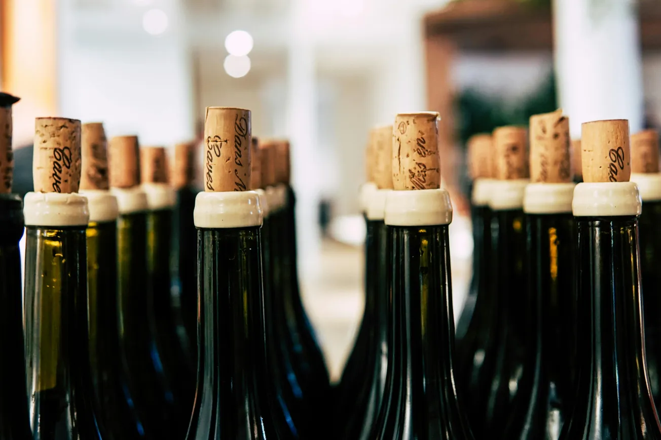 Rows of green wine bottles with corks, closely packed, creating a pattern. Soft, blurred background suggests a warm, inviting atmosphere.